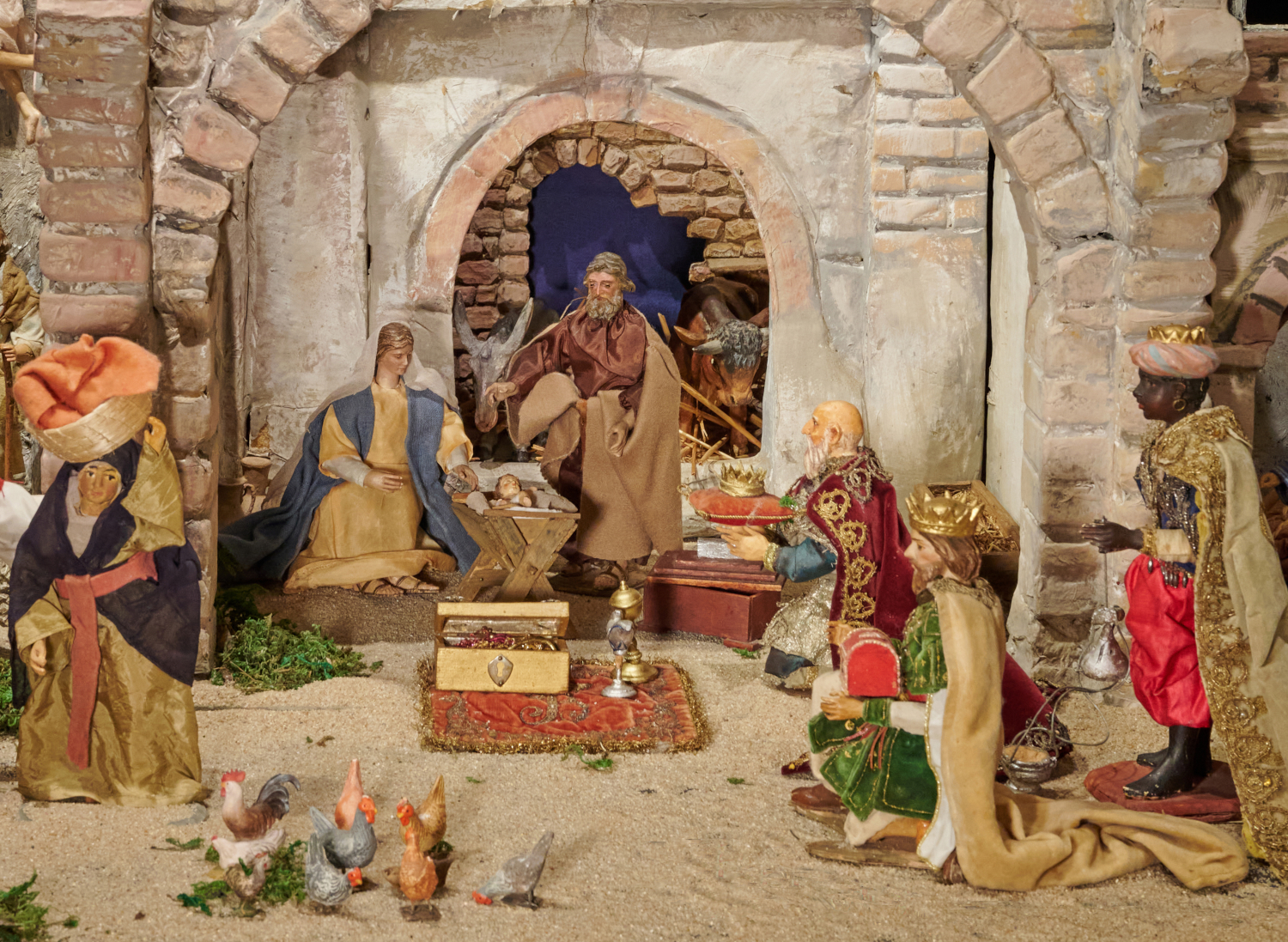 Blick in Detail einer größeren Weihnachtskrippe auf die Szene der Heiligen Familie und die Anbetung der Heiligen Drei Könige. Die Heilige Familie befndet sich in Frontalansicht in einem gemauerten Stall. Maria sitzend links, Kind in Krippe mittig und Josef stehend rechts. Davor sind die Gaben der Könige auf einem Teppich ausgebreitet. Die Heiligen Drei Könige sind auf der rechten Bildseite von der Seite zu sehen. Ganz rechts an Bildrand steht Melchior, Caspar und Balthasar hingegen knien. Am linken Bildrand steht eine Frau mit einem großen Korb auf dem Kopf tragend. Im Vordergrund einige Hennen.