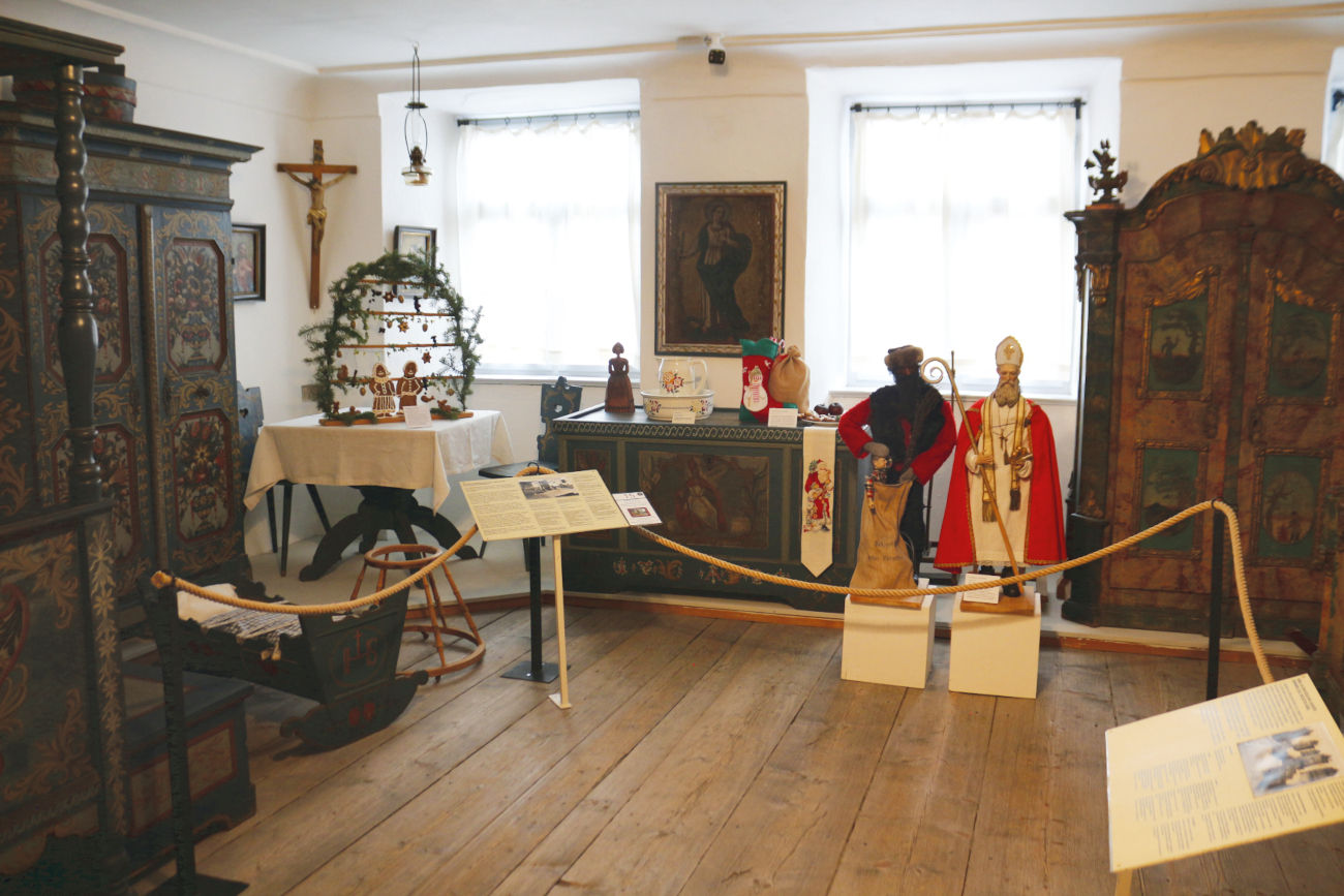 Blick in einen Raum der Dauerausstellung mit Bauernmöbeln in Richtung Fenster. Zu sehen sind von links nach rechts: Teile eines Himmelbetts, Kinderwiege, Schrank, Tisch, Truhe in Bildmitte und Schrank. An den Wänden sind Bilder und im linken Eck ein Kruzifix angebracht. Auf dem Tisch steht ein sogenannter Paradiesbaum, ein alter weihnachtlicher Schmuck. Er stellt einen Bogen dar, der mit Immergrün und kleinen Anhängern verziert ist. Zwischen der Truhe und dem rechten Schrank stehen zwei Figuren auf weißen Sockeln: links der Krampus im schwarzen Gewand und rechts der Nikolaus mit weißem Gewand und rotem Mantel.