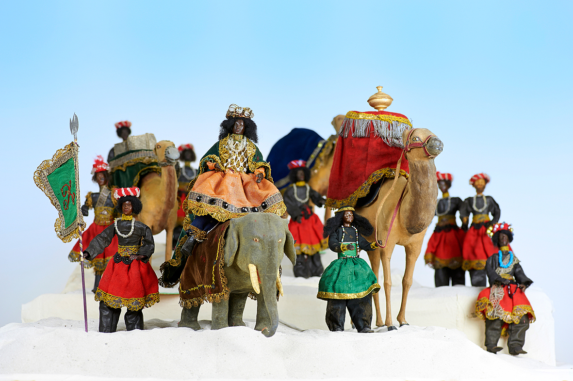 Vor hellblauem Hintergrund auf einem weißen hügeligen Untergrund steht eine Gruppe von Krippenfiguren. Es handelt sich um den Königszug von einem der Heiligen Drei Könige. Im Vordergrund mittig sitzt er auf einem Elefanten. Er trägt ein prächtiges Gewand in Blau, Grün und Orange mit goldenen Spitzenborten und eine Krone auf dem Kopf. Links wird der Elefant von einem Begleiter mit grüner Fahne flankiert. Rechts vorne steht ein Kamel mit hohem roten Podest auf dem Rücken tragend, auf dem ein goldenes Gefäß steht. Die Zügel des Tiers werden von einem Begleiter zu Fuß gehalten. Dahinter sind weitere Kamele mit Begleiter zu sehen. Alle, bis auf den vorderen Kamelführer in Schwarz-Grün, tragen ein ähnliches Kostüm in Schwarz-Rot und Perlenketten um den Hals. 