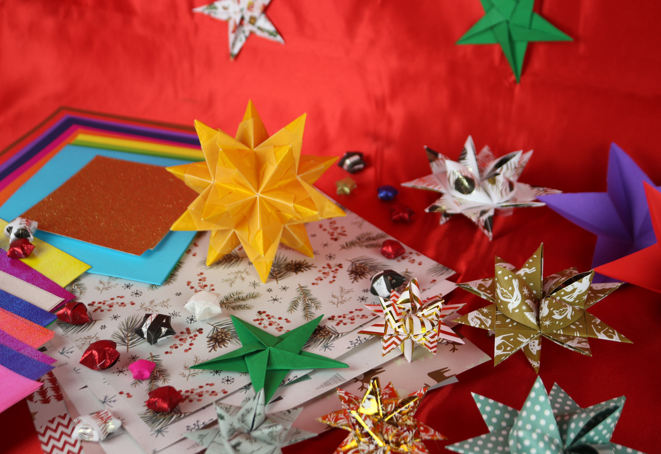 Vor rotem Hintergrund befinden sich mehrere aus Papier gebastelte bunte, gemusterte und glitzernde Sterne, außerdem Papierbögen als Bastelmaterial.