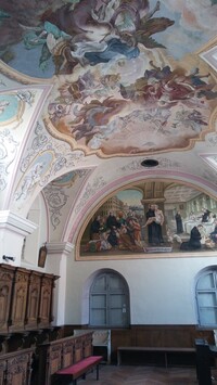 Foto eines sakralen Raumes mit bemalter Stuckdecke und -wand sowie hölzernem Gestühl.