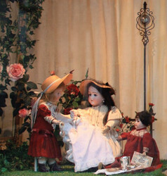 Blick in ein Schaufenster. Arrangement von drei historischen Puppen auf einer grünen Wiese, im Hintergrund Rosenranken und -blüten sowie ein weißer Vorhang. Mittig eine Puppe mit weißem Kleid, dunklen langen Haaren und einem Strohhut. Links stehende Puppe mit rotem Kleid, blonden schulterlangen Haaren und einem Strohhut mit weißer Borte. Rechts sitzende kleinere Puppe mit rotem Gewand, kurzen dunklen Haaren und einem Blatt Papier in der Hand. Zudem liegen neben ihr einige Gegenstände auf der Wiese. Im Hintergrund rechts eine dekorative Blumenstange. 