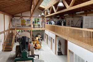 Innenansicht einer Ausstellungsfläche. Blick von einer Galerie im Obergeschoss in einen großen hallenartigen Raum mit Deckenkonstruktion aus Holz und Glas. Im Erdgeschoss sind mehrere große Fahrzeuge verteilt, die teilweise über Walzen und lokartige Schornsteine verfügen. Zudem sind im Hintergrund auf einer umlaufenden Empore mit Holzgeländer und Treppe an den Wänden verschiedene Gegenstände angebracht, vor denen eine größere Personengruppe steht.