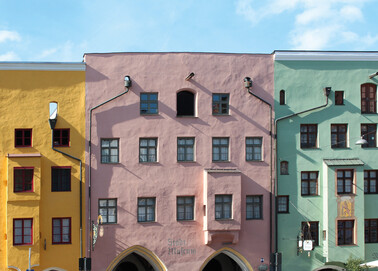 Ansicht der oberen Fassadenstockwerke von drei nebeneinander liegenden, Baustil ähnlichen Häusern mit flachen Dachabschlüssen und Erkern. Das mittlere Haus ist rötlich und in voller Breite abgebildet. Das linke als auch rechte Gebäude sind angeschnitten. Das linke Haus ist in kräftigem Gelb und das rechte Haus in einem helleren Grünton gehalten. Oben ist ein blauer Streifen Himmel erkennbar.