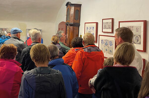 Versammlung einer Personengruppe vor einer Wand mit mehreren aufgehängten Bildern und einer Standuhr. Eine Person in der Mitte zeigt erklärend zu den Objekten.