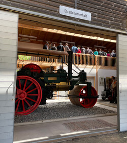 Fotografie: Blick durch ein geöffnetes Tor auf eine große Walze, die an eine historische Lokomotive erinnert. Mittig über dem Tor ist ein Schild mit Aufschrift "Straßenunterhalt". Im Hintergrund steht auf einer Galerie eine Personengruppe.