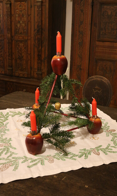 Blick von oben auf einen alten Holztisch mit Tischdecke, auf der um den Rand grüne Zweige aufgestickt sind. Darauf steht ein Weihnachtsschmuck, ein sogenanntes Paradeisl. Dieses besteht aus vier Äpfeln, die mit Zweigen miteinander verbunden sind. Dabei bilden drei Äpfel ein Dreieck als Basis. Der vierte Apfel ragt oben in der Mitte, darunter hängt eine goldene Walnuss. Auf allen vier Äpfeln sind rote Kerzen angebracht. Zudem sind grüne Tannenzweige in die Konstruktion gesteckt. Im Hintergrund sind Teile von mit dunklen Ornamenten bemalten Holzschränken erkennbar.