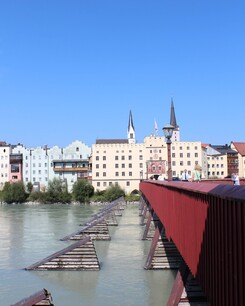 Foto einer frontalen Längsansicht eines roten Brückengeländers mit Fluss, zulaufend auf eine Häuserfront.
