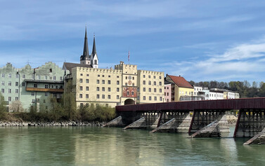 Blick auf eine bunte Fassadenfront bestehend aus mehreren aneinander liegenden Häusern an einem Fluss vom gegenüberliegenden Ufer aus fotografiert. Über den Fluss verläuft eine rote Brücke mit Eisbrechern an den Pfeilern zu einem größeren Gebäudeensemble zu. In dessen Mitte befindet sich ein turmartiges Gebäude mit Tordurchfahrt, teilweise Fassadenbemalung, Dachzinnen und Fahne. Auch die beiden größeren Gebäude links und rechts des Torturms verfügen über Dachzinnen, zudem über vertikal wie horizontal parallel verlaufende Fenster. Im Hintergrund sind Kirchtürme erkennbar. 