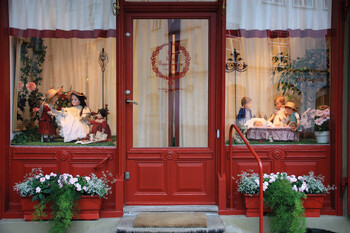 Blick auf eine Schaufensterfront mit mittiger roter Glastüre, die von je einem größeren Fenster mit roter Umrahmung flankiert wird. Auf der Türe befindet sich ein rotes rundes Logo. Vor den Fenstern steht je ein üppig bepflanzter roter Blumenkasten. In den Schaufenstern sind Szenerien mit mehreren Puppen arrangiert. Der hinter der Glasfront befindliche Innenraum wird durch einen weißen Vorhang mit roter Borte verdeckt. 