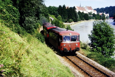 Foto eines roten Zuges, der in einer hügeligen Landschaft fährt. Rechts ist ein Fluss und im Hintergrund Häuser einer Stadt erkennbar.