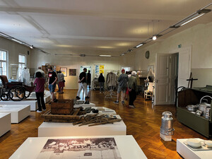 Blick in einen größeren Ausstellungsraum mit Parkettboden. Darin sind links und vorne mittig mehrere weiße niedrige Blöcke verteilt, auf denen diverse Gegenstände, wie zum Beispiel ein alter Rollstuhl, präsentiert werden. Außerdem sind Ständer mit Kleidung und weitere Objekte ausgestellt. Im Hintergrund sehen sich mehrere Personen in der Ausstellung um.