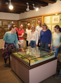 Versammlung einer Personengruppe um eine flache, hüfthohe Vitrine in einem Raum mit hölzerner Balkendecke. In Vitrine ist ein Modell einer Landschaft bzw. Stadt erkennbar. Im Hintergrund an den beiden sichtbaren Wänden hängen mehrere Bilder hinter flachen Glasvitrinen.