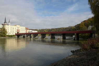 Foto einer roten Brücke, die über einen Fluss verläuft. Links mündet sie in eine größere Gebäudefront.
